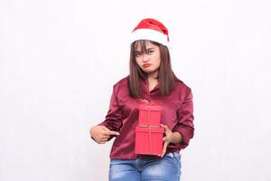 beautiful young asian indonesian girl pouting carrying gift box in christmas santa claus hat modern red shirt outfit pointed side box on white background for promotion and advertising photo