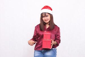 beautiful young asian indonesian girl joyfully carrying gift box in christmas santa claus hat modern red shirt outfit pointed side box on white background for promotion and advertising photo