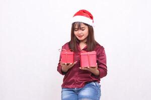 Photo portrait of young beautiful asian girl carrying gift box in christmas santa claus hat modern shiny red shirt outfit hands raise and look at box on white background for promotion and advertising