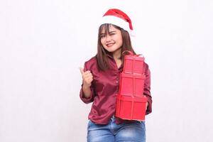 beautiful young southeast asian girl happy carrying 3 boxes of gifts at christmas wearing santa claus hat modern red shirt outfit thumbs up sign okay all white background for promotion and advertising photo