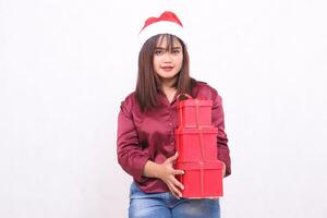 Cheerful young beautiful Southeast Asian girl carrying 3 boxes of gifts at Christmas wearing Santa Claus hat modern red shirt outfit holding all white background for promotion and advertising photo
