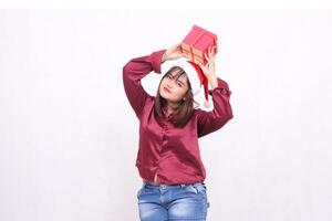 Portrait photo of beautiful plain aged Asian girl carrying gift box in Christmas Santa Claus hat modern shiny red shirt clothes lifting box on white background for promotion and advertising