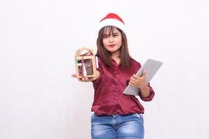 beautiful young southeast asian woman smile give gift food package in hamper and laptop tablet at christmas wearing santa hat wearing red shirt modern white background for promotion and advertising photo