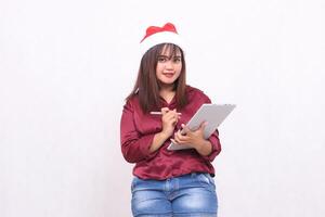 beautiful young southeast asian woman writing order looking at camera while carrying laptop tablet at christmas wearing santa hat wearing red shirt modern white background for promotion photo