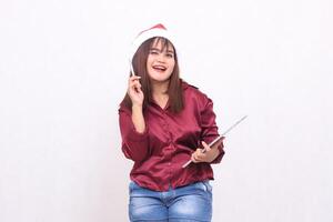 Cheerful young beautiful Southeast Asian woman holding up pen while carrying laptop tablet at Christmas wearing Santa hat wearing red shirt modern white background for promotion and advertising photo