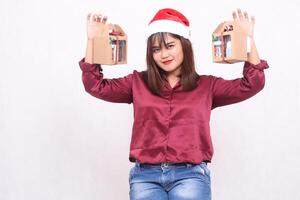 Beautiful young Southeast Asian woman smiling hands raised carrying 2 boxes of hamper gifts at Christmas wearing Santa Claus hat modern red shirt outfit white background for promotion and advertising photo