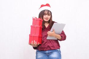 hermosa joven Sureste asiático mujer sonriente que lleva 3 regalo cajas cesto y mirando a ordenador portátil tableta a Navidad vistiendo Papa Noel sombrero moderno rojo camisa atuendo blanco antecedentes para promoción foto