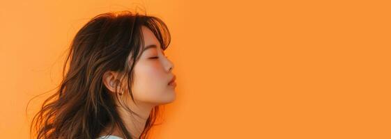 A woman with black hair stands in front of an orange wall, eyes closed, smiling photo