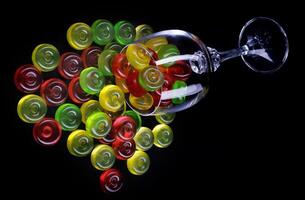 A lot of candy canes are scattered from an overturned glass goblet. Multi-colored candies in a glass on a black background. photo