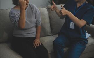 Smiling kind pleasant young female doctor in white uniform visiting disabled elderly mature patient at home, asking about health condition at checkup meeting, geriatric healthcare help concept. photo