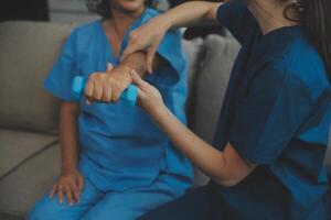 Old woman training with physiotherapist using dumbbells at home. Therapist assisting senior woman with exercises in nursing home. Elderly patient using dumbbells with outstretched arms. photo