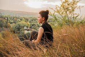 un contemplativo mujer se sienta en medio de alto césped, mirando a un sereno paisaje durante puesta de sol foto