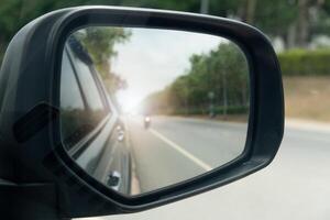 Inside view of mirrors wing. Rear view of a gray car with asphalt road and green trees in the daytime. Blurred image of a motorcycle approaching from behind. photo