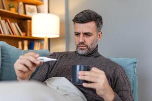 Man feeling sick lying in the bed and looking the thermometer. Sick man lying on sofa checking his temperature under a blanket at home in the living room photo