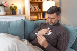 Man feeling sick lying in the bed and looking the thermometer. Sick man lying on sofa checking his temperature under a blanket at home in the living room photo