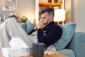 Sick sad man sits on couch at home suffers from runny nose flu disease coronavirus pandemic covid epidemic sneezes. Unwell guy feeling bad fever virus illness symptoms indoor photo