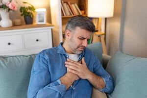 Man presses hand to chest has heart attack suffers from unbearable pain, Mature man with pain on heart in living room. man suffering from bad pain in his chest heart attack at home photo