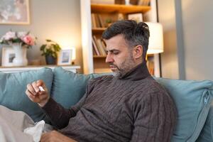 Man feeling sick lying in the bed and looking the thermometer. Sick man lying on sofa checking his temperature under a blanket at home in the living room photo