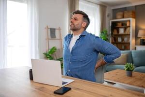 Shot of a businessman suffering from a backache while working at his desk in his office. Man having back pain while working on laptop from home photo