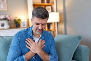 Man presses hand to chest has heart attack suffers from unbearable pain, Mature man with pain on heart in living room. man suffering from bad pain in his chest heart attack at home photo