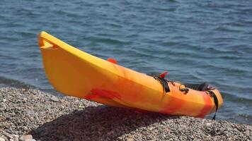 un' giallo e arancia kayak è seduta su un' roccioso spiaggia. il kayak è non nel uso e è pendente contro il rocce. il scena è tranquillo, calmo e sereno, con il calma acqua. video