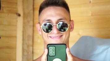 The young man in sunglasses, sunbathing by the sea and spending time on his mobile phone texting with girlfriend, looks at the camera in surprise, sees a news on the phone. video