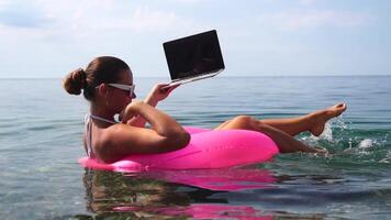 mujer persona de libre dedicación trabajos en ordenador portátil nadando en mar en rosado inflable anillo. contento turista en Gafas de sol flotante en inflable rosquilla y trabajando en ordenador portátil computadora en calma océano. remoto trabajando en cualquier sitio video