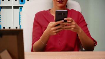 Woman browsing on smartphone smiling sitting on desk in modern office room. Hispanic entrepreneur working in professional workplace in corporate company typing on mobile phone in front of computer video