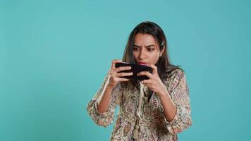 Gamer with disappointed look on face after receiving game over screen, holding smartphone. Indian woman upset after losing videogame, playing with phone, isolated over studio background video