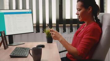 Woman worker writing on mobile phone in front of computer in office room. Hispanic entrepreneur sitting in corporate company workplace, taking break typing on smartphone in front of pc reading news video