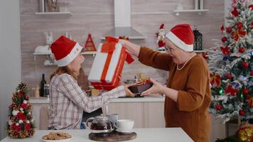 Grandmother suprising grandchild with wrapper present gift enjoying traditional winter holiday together in decorated xmas kitchen. Happy family with santa claus hat celebrating christmas season video