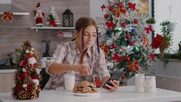 Porträt von Kinder Sitzung beim Tabelle im Weihnachten dekoriert Küche Surfen auf Sozial Medien mit Smartphone. kaukasisch Mädchen genießen Weihnachten Urlaub Essen traditionell hausgemacht Kekse Trinken Milch video