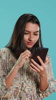 Vertical Woman happily scrolling on phone touchscreen to check social media feed, amazed by photo. Indian person browsing internet websites, astonished by content, studio background, camera A video