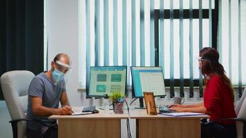 People with mask and visor working in new normal office checking reports and writing on computer. Coworkers in modern workplace respecting protection rules against covid virus using plexiglass. video