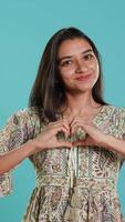 Vertical Portrait of jolly friendly indian woman doing heart symbol shape gesture with hands. Cheerful nurturing person showing love gesturing, isolated over studio background, camera A video