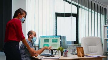 Colleagues with masks discussing graphics sitting in new normal office room. Coworkers talking working pointing at desktop respecting social distancing against covid virus using plexiglass. video