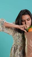 Vertical Indian woman with ecological paper bag in hands looking at bell pepper, living healthy lifestyle. Ecology lover holding shopping bag with organic produce, studio backdrop, camera A video