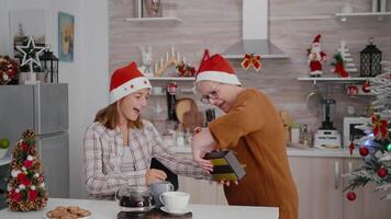Grandmother surprising grandchild with xmas wrapper gift present celebrating christmastime in decorated kitchen. Happy family enjoying christmas holiday during winter season video