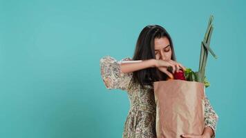 vrouw met papier zak met groenten testen kwaliteit, op zoek Bij komkommer, geïsoleerd over- studio achtergrond. persoon inspecteren boodschappen na buying hen van nul verspilling winkel, camera een video