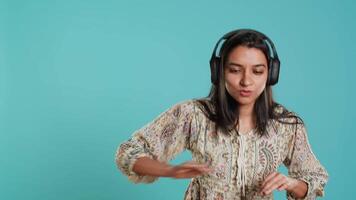 Happy woman playing electronic music and waving hand in air, pretending to use turntables. Cheerful DJ wearing headphones, producing beats, making songs, isolated over studio background, camera A video
