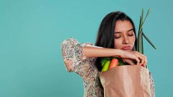 femme avec papier sac dans mains rempli avec des légumes vérification carotte pour impuretés. conscient vivant client avec acheté les courses à être utilisé comme cuisine ingrédients, studio arrière-plan, caméra b video