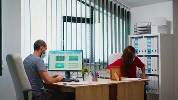 Hispanic woman with mask helping colleague with reports sitting in office room in front of computer. Coworkers discussing working in new normal workplace typing on pc keyboard pointing at desktop video