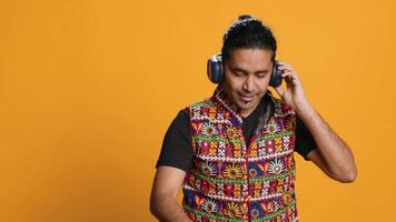 Indian man playing music, having fun moving on rhythm, pretending to use turntables. Upbeat DJ wearing headphones, producing beats, isolated over studio background, camera A video