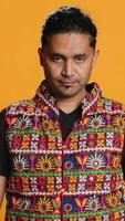 Vertical Portrait of optimistic indian man wearing colorful vest with positive facial expression smiling, looking pleased. Calm person in traditional clothing, feeling satisfied, studio backdrop, camera A video