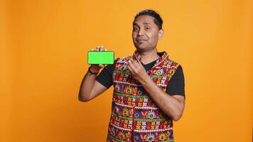 Content creator presenting green screen mobile phone, isolated over studio background. Indian person reviewing copy space mockup smartphone device for internet channel, camera B video