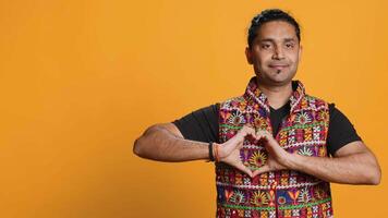 Portrait of jolly friendly indian man doing heart symbol shape gesture with hands. Cheerful nurturing person showing love gesturing, isolated over studio background, camera A video