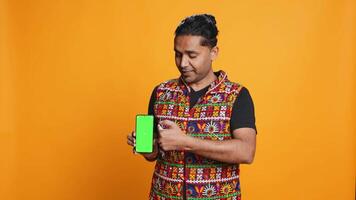 Influencer presenting green screen mobile phone, isolated over studio background. Indian person holding copy space chroma key smartphone used for advertising brands, camera B video