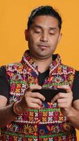 Vertical Cheerful Indian person in traditional clothing entertained by videogames on smartphone. Joyous player enjoying game on phone, having fun online, studio backdrop, camera A video