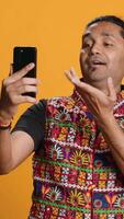 Vertical Joyous Indian man greeting friend over online videocall on cellphone, doing friendly gestures. Excited person bonding with mate using mobile phone videoconference, studio background, camera A video