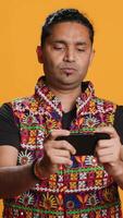 vertical contento indio hombre entretenido por videojuegos en teléfono inteligente, disfrutando ocio tiempo. jugador disfrutando juego en teléfono, teniendo divertido derrotando enemigos, aislado terminado estudio fondo, cámara un video